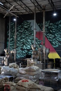 Inside Black and White Coffee's roasting facility, stacks of raw coffee beans sit in the foreground. The back wall features a mural with leaves and a gradient. In front of the mural sit coffee roasting machines. 