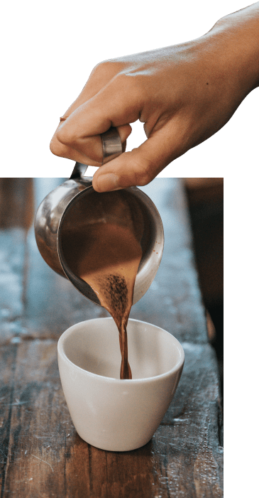 A hand pours espresso from a stainless steel pitcher into a white porcelain mug.