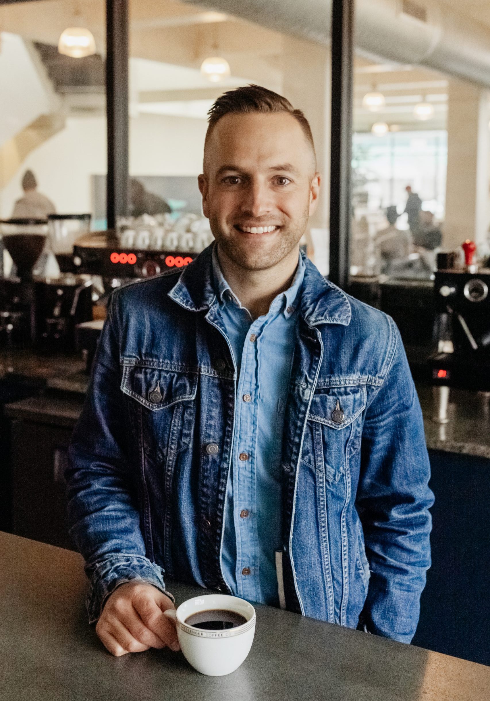 A white man stands in front of an espresso machine in a cafe setting. He's smiling and wearing a light blue button-up shirt under a denim jacket.