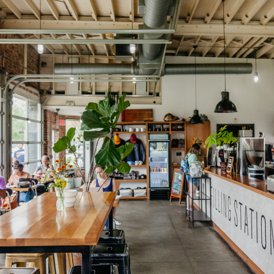The interior of a Filling Station café location. The space features concrete floors and a coffee bar displaying the Filling Station logo. Bright light filters into the space filled with tables, stools, and live plants.